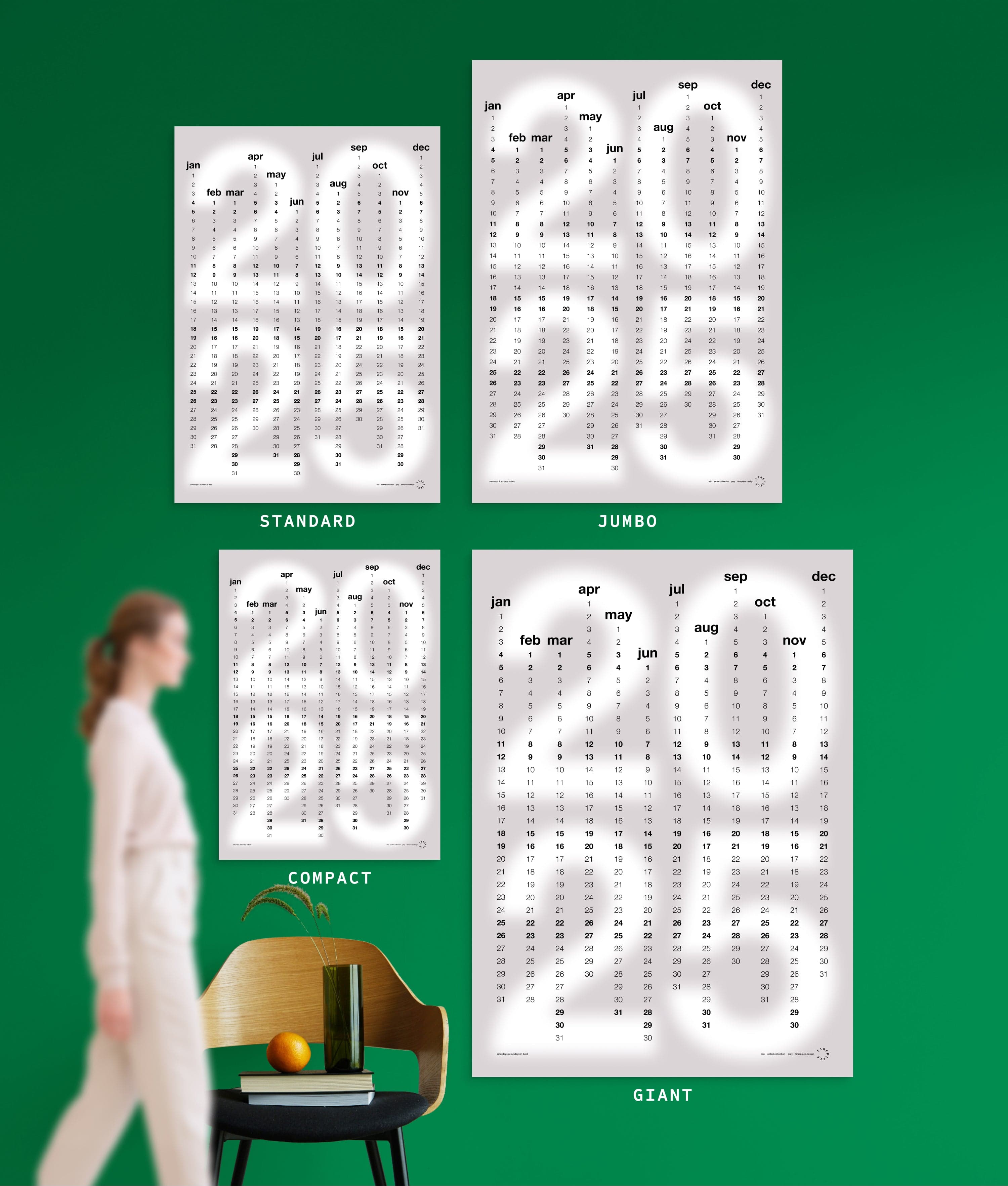 4 sizes of Min in Grey colour yearly planner on green wall, with person, chair, orange and vase shown for scale